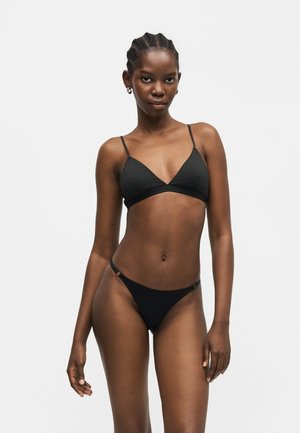 Mujer delgada con el cabello trenzado, vistiendo un conjunto de bikini negro de triángulo, de pie frente a un fondo liso gris claro, mirando hacia adelante.