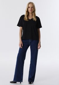 Zwarte T-shirt met korte mouwen en V-hals, gecombineerd met wijde blauwe denim jeans en zwarte puntige schoenen. Vlotte stof en relaxte pasvorm.