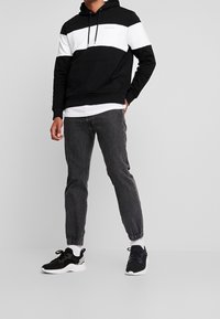 Felpa con cappuccio bianca e nera con tasca frontale e logo, abbinata a jeans grigi e sneakers nere con suole e dettagli bianchi.