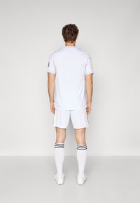 Maglietta atletica bianca a maniche corte, abbinata a pantaloni corti bianchi e calze a righe nere. Accenti gialli sui pantaloni corti, tessuto leggero.