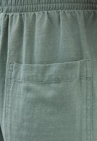 Groene stof met een lichte glans, voorzien van een geplooide elastische tailleband en een enkele opgestikte zak aan de voorkant. Textuurdetail op het oppervlak zichtbaar.