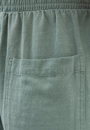 Zelená tkanina se slabým leskem, s nařaseným elastickým pasem a jednou nášivkou na přední straně. Viditelné texturované detaily na povrchu.