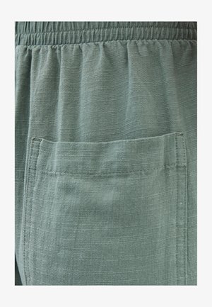 Grønne stof med en let glans, med en rynket elastisk talje og en enkelt påsyet lomme foran. Teksturdetaljer synlige på overfladen.