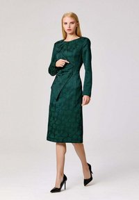 Robe imprimée florale vert foncé, à manches longues et design ajusté. Dotée d'une ceinture à la taille et d'un ourlet au genou. Associée à des talons noirs.