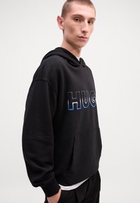 Svart hettegenser med frontlomme og "HUG"-tekst i blå gradient. Laget av myk stoff med ribbestrikkede mansjetter og hette.