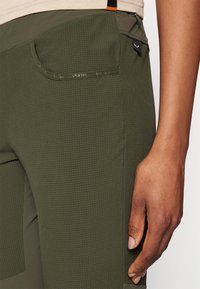 Gröna byxor med en texturerad yta, med en sidoficka och en figurnära design. Materialet verkar lätt och hållbart.