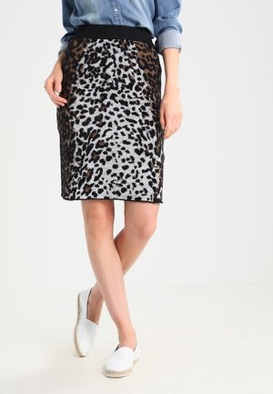 Vrouw draagt een knielange rok met luipaardprint, een opgerolde spijkershirt en witte instapschoenen, staand met gekruiste benen.