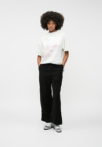 Fehér grafikás póló rózsaszín nyomattal, fekete bőszárú nadrággal és ezüst cipővel párosítva. A modell a zsebében lévő kezekkel áll egy egyszínű háttér előtt.