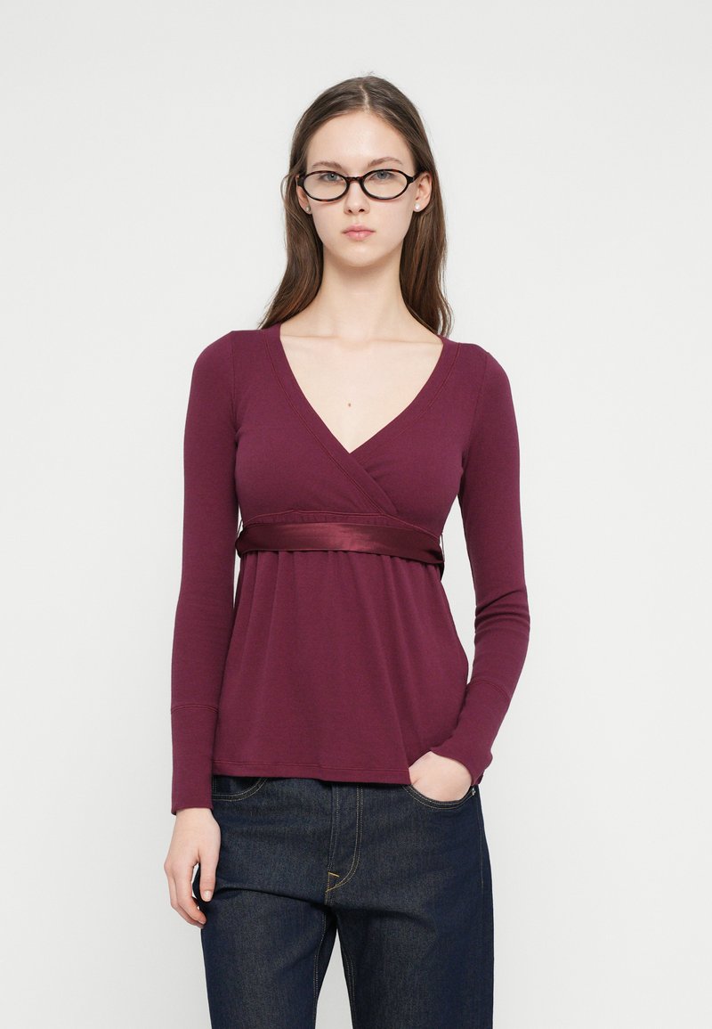 Νεαρή γυναίκα με μακριά καστανά μαλλιά και γυαλιά, που φοράει μακρυμάνικη μπλούζα σε σκούρο μοβ χρώμα τύπου wrap και σκούρα μπλε τζιν, στέκεται μπροστά σε απλό φόντο.