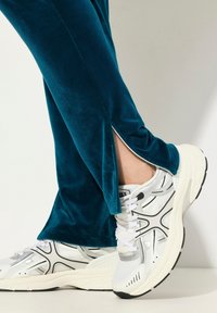 Teal velours broek met zijsplitdetails, gecombineerd met witte, zwart-geaccentueerde sportschoenen van mesh en synthetische materialen.