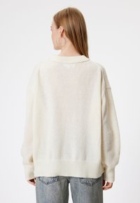 Przesadny sweter w kremowym kolorze z prążkowanym wykończeniem u dołu i na rękawach, cechujący się miękką dzianiną i subtelnym wywiniętym kołnierzem. W zestawie z jasnoniebieskimi dżinsami.