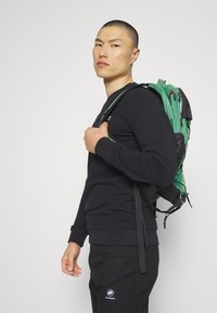 Zaino verde e nero con molteplici scomparti, spallacci regolabili e dettagli hardware frontali, indossato da una persona in un felpa nera.