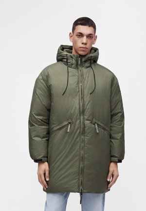 Jonge man met een olijfgroene pufferjas met capuchon, zwarte trekkoorden en met rits afsluitbare zakken, staand tegen een effen witte achtergrond.