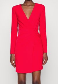 Kvinna klädd i en figurnära, långärmad, klar röd klänning med djup V-ringning och draperad detalj vid midjan, stående mot en enkel bakgrund.