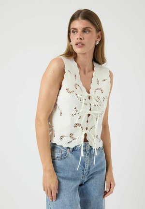 Hvid blonde ærmeløs top med bølgede kanter, udskårne blomsterdesigns og en frontknude, parret med lyseblå jeans.