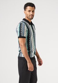 Pletené polo tričko s alternujícími vzory modré, zelené a bílé barvy, vybavené černým límečkem a žebrovaným lemem. Krátké rukávy, přiléhavý střih.