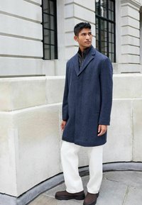 Dunkelblauer Wollmantel mit Reverskragen, kombiniert mit einer schwarzen Zip-Jacke, weißen Hosen und braunen Stiefeln, die vor einer Steinmauer steht.