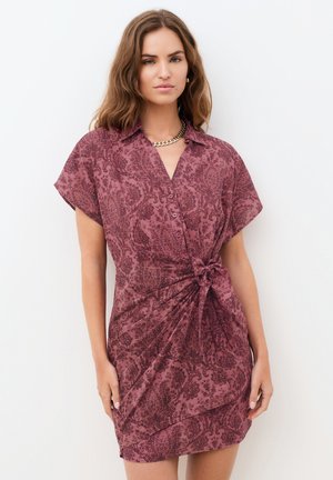 Nainen aaltoilevine ruskeine hiuksineen, jolla on päällään lyhythihainen, napitettu viininpunainen mekko, jossa on kukkakuosi ja sivusolmu, seisomassa valkoista taustaa vasten.