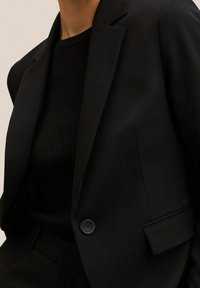 Černé přizpůsobené sako s jedním knoflíkem, ostrými klopami a strukturovaným siluetou, doplněné o černý svetr s žebrováním.