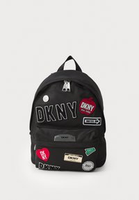 Black fabric backpack featuring multiple patches, zippers, and a handle. Notable patches include "DKNY" and various logos. Compact design.