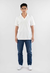 Polo bianco con scollo a V, maniche corte, abbinato a jeans in denim blu e sneakers bianche. Design semplice, senza motivi o dettagli visibili.