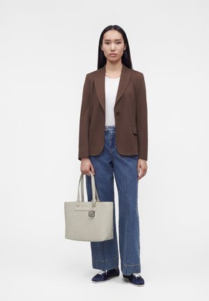 Nainen ruskeassa bleiserissä, valkoinen toppi, leveälahkeiset siniset farkut, tummansiniset lenkkarit, pitää beigeä kohokuvioitua olkalaukkua.