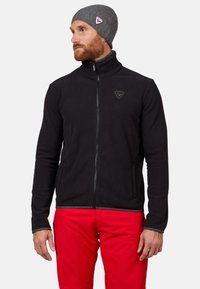 Schwarze Fleecejacke mit hohem Kragen, Frontreißverschluss und Logodetail. Getragen mit roten Hosen und einer grauen Strickmütze. Glatte Textur und taillierte Passform.