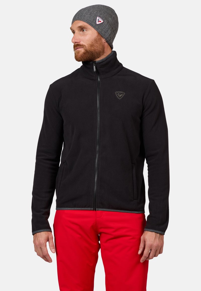 Schwarze Fleecejacke mit hohem Kragen, Frontreißverschluss und Logodetail. Getragen mit roten Hosen und einer grauen Strickmütze. Glatte Textur und taillierte Passform.