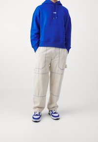 Felpa blu con tasca frontale e cappuccio con lacci, abbinata a pantaloni beige chiaro con cuciture a contrasto e una vestibilità rilassata, completata da sneakers blu e bianche.