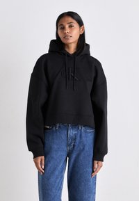 Felpa nera cropped con logo frontale, maniche lunghe e cappuccio con cordino, abbinata a jeans blu larghi. Design semplice e casual.