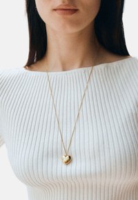 Colgante dorado en forma de coraz�ón con una cadena fina, llevado sobre una blusa blanca con texturas. El colgante es liso y refleja la luz. Diseño simple y elegante.