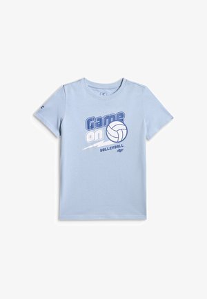 Garçon debout avec un t-shirt bleu clair à thème volley-ball, un short cargo vert, des chaussettes blanches, des baskets, et tenant un sweat gris.