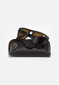 Ochelari de soare Ray-Ban cu lentile de culoare maro, în rame dintr-un tortoise dark, așezați pe un capac texturat de culoare neagră cu un logo aurit și închidere tip snap.