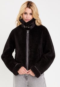 Schwarze, flauschige Jacke mit hohem Kragen, Reißverschluss und kontrastierenden schwarzen Lederakzenten am Kragen. Sie hat weite Ärmel und einen strukturierten Stoff.