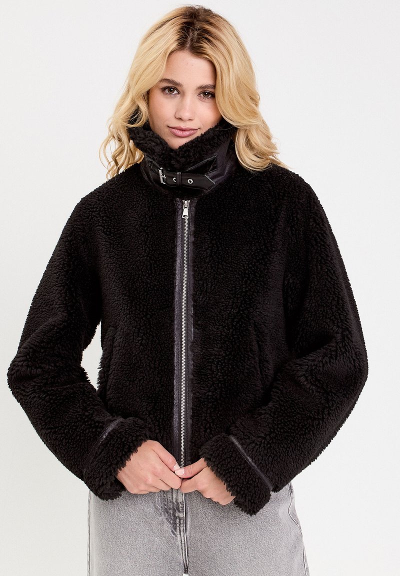 Schwarze, flauschige Jacke mit hohem Kragen, Reißverschluss und kontrastierenden schwarzen Lederakzenten am Kragen. Sie hat weite Ärmel und einen strukturierten Stoff.