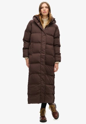 Bruine pufferjas met een lange, gewatteerde ontwerp, capuchon en een rits aan de voorkant. Combinatie met bruine veterschoenen met gele veters.