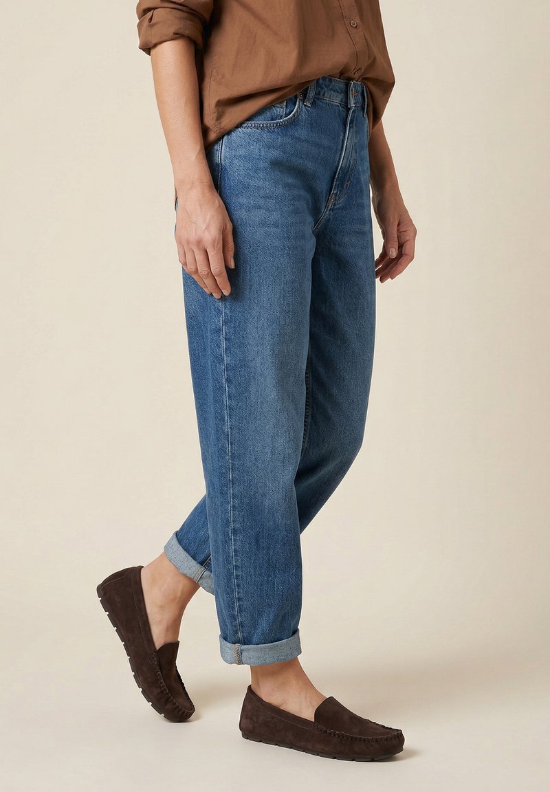 Person, die blaue umgeschlagene Jeans, braune Wildleder-Mokassins und ein hochgekrempeltes braunes Hemd trägt und vor einem schlichten Hintergrund steht.