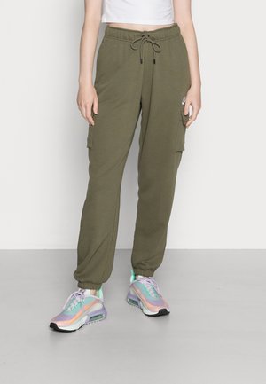 Femme portant un jogger cargo vert olive à taille élastique avec cordon de serrage et des baskets multicolores iridescentes, debout sur un sol gris.