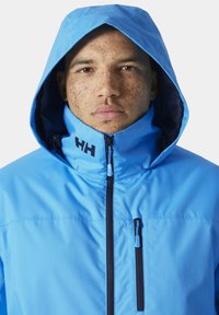 Lichtblauwe waterdichte jas met een capuchon, een ritssluiting aan de voorkant en contrasterende marineblauwe accenten. Heeft een groot logo op de nek.