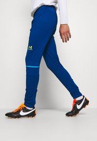 Modré sportovní kalhoty vyrobené z lehkého materiálu s elastickým pasem, s modrým akcentem a logem Under Armour.