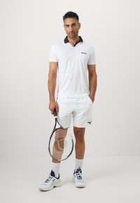Weißes Polohemd mit schwarzem Kragen, Logo auf der Brust, kombiniert mit weißen Shorts. Hält einen Tennisschläger, trägt Sportschuhe mit blauen Akzenten.