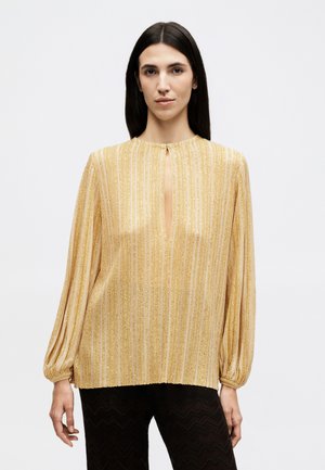 Γυναίκα με μακριά σκούρα μαλλιά φοράει χρυσό μεταλλικό ριγωτό μπλουζάκι με λαιμόκοψη σε σχήμα κλειδαρότρυπας και μαύρο παντελόνι με υφή, στέκεται μπροστά σε λευκό φόντο.