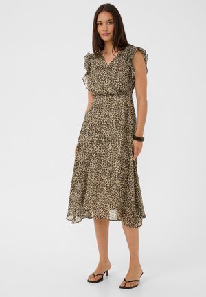 Femme aux longs cheveux foncés portée d'une robe midi sans manches à imprimé léopard et de sandales noires à talons, debout devant un fond blanc.