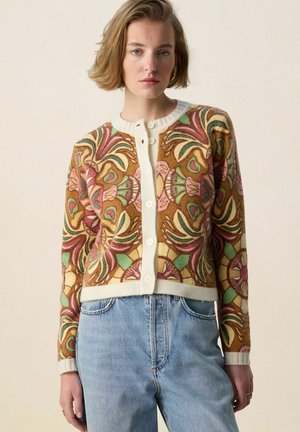 Femme portant un cardigan boutonné avec des motifs floraux multicolores et un jean taille haute bleu clair, debout devant un fond beige uni.