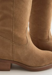 Bēšas zamša zābaki līdz ceļiem ar sakrautu koka papēdi. Raksturīgas izliektas šuves un gluda tekstūra, kas uzsver elegantus dizaina elementus.