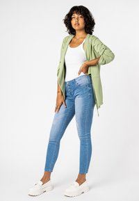 Lichtgroene gebreide cardigan over een witte tanktop, gecombineerd met blauwe skinny jeans met een knoopband en witte platform schoenen.