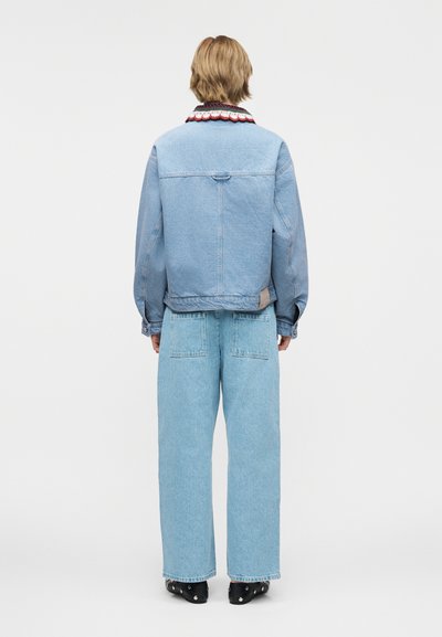 Άτομο με κοντά ξανθά μαλλιά που φοράει ένα ανοιχτό γαλάζιο τζιν μπουφάν και φαρδιά τζιν, στρέφεται με την πλάτη προς το απλό φόντο.