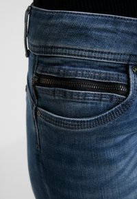 Farkut keskipitkässä sinisessä sävyssä, joissa on vetoketjullinen takatasku, hienovaraisia ompeluyksityiskohtia ja haalistunut rakenne kankaassa.
