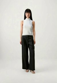 White sleeveless turtleneck top paired with high-waisted black trousers. Model wears light-colored heeled sandals. Simple, clean design.
