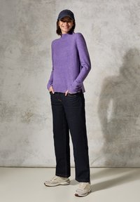 Lila Strickpullover mit hohem Kragen, kombiniert mit dunklen Jeans und weißen Sneakers. Eine marineblaue Mütze rundet das Outfit ab.